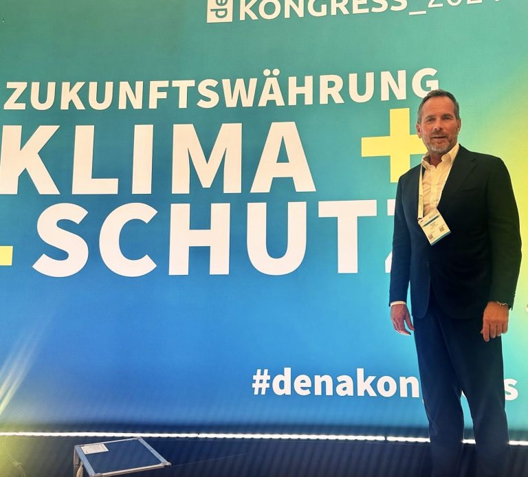 dena Klimakongress Mann steht vor einem großen Banner mit dem Text „Zukunftswährung Klima+Schutz“.